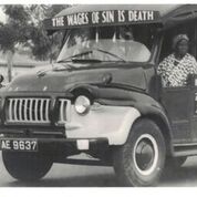 A trader sitting in the front seat of a mammy truck with the inscription “The Wages of Sin is Death” -  “Lorry Slogans,” <em>Mate Wayo</em>, 2 March 1971. Source: PS 402/12 Photographic Section, Information Services Department, Ministry of Information, Accra, Ghana.” width=”50%” height=”auto”>

<figcaption><small>Figure 2-a. A trader sitting in the front seat of a mammy truck with the inscription “The Wages of Sin is Death” – “Lorry Slogans,” <em>Mate Wayo</em>, 2 March 1971. Source: PS 402/12 Photographic Section, Information Services Department, Ministry of Information, Accra, Ghana. Rightsholder: Ministry of Information, Accra, Ghana.</small></figcaption></figure>
<h3>Training drivers</h3>
Drivers worked hard in and on their vehicles, developing wide-ranging skill and expertise. Driving was an implicitly male profession. Drivers defined the physical strength necessary to operate vehicles in poor road conditions and load goods, as well as the social and emotional control necessary to engage with passengers, as male traits even as they established formal business arrangements with female passengers or drove for female vehicle owners. Young men were trained through apprenticeships with master drivers. Families paid fees (often in some combination of alcohol, cigarettes, and cash) to the master, who promised to train the young man. Apprentices (or “mates”) often lived in the master’s household and traveled with them constantly. Daily duties included washing the vehicle, loading and unloading goods, and recruiting passengers. Throughout their daily work, mates closely observed their master’s behavior to learn both the tangible, practical skills of driving and vehicular maintenance but also the more intangible social skills and cultural knowledge necessary for a successful driving career. This rigorous training often lasted 5 years or more, producing highly qualified and professional drivers who possessed not only the technical expertise necessary for driving work, but who were also integrated into a tight-knit community of drivers, defined by high levels of professionalism and, beginning in the mid-1930s, organized into unions.
<figure><a href=